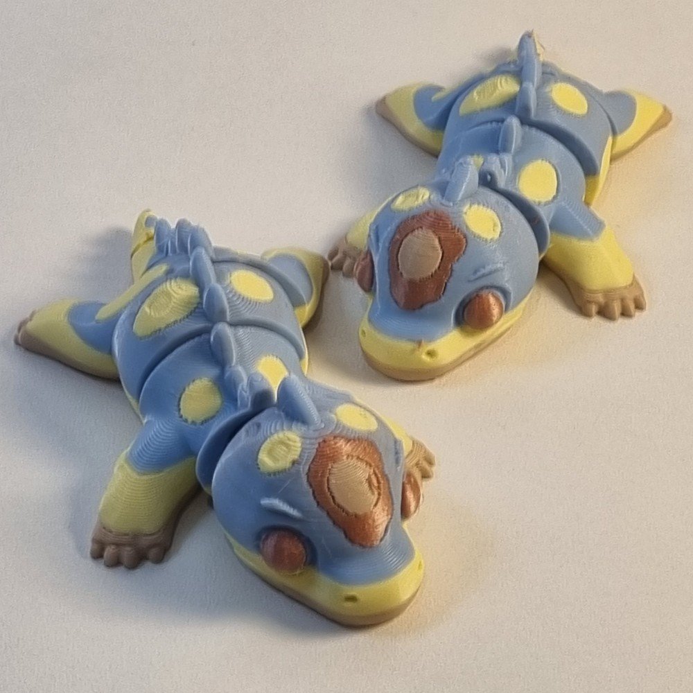 Dois brinquedos dinossauro articulado fidget toy impressos em 3D nas cores azul, amarelo e marrom, deitados lado a lado sob fundo claro.